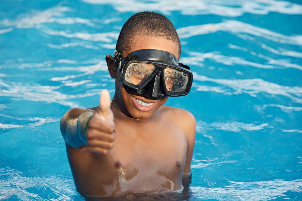 A smiling boy in a swimming pool gives a thumbs-up while wearing snorkeling goggles, showcasing enjoyment in a fun aquatic environment.