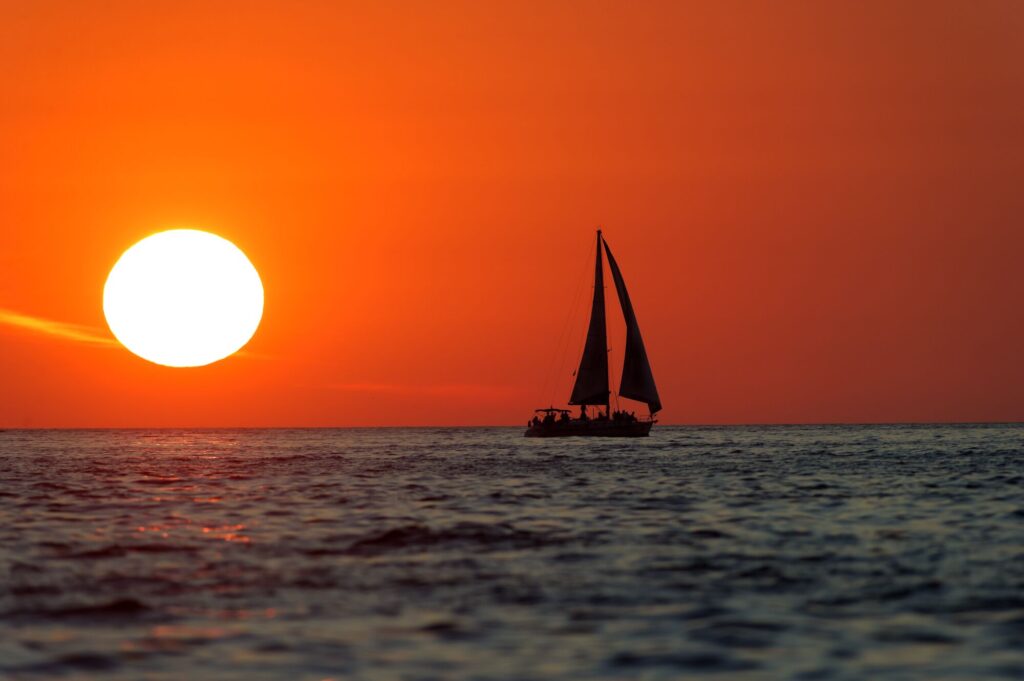 Sailboat glides across calm waters at sunset, with a large, bright sun casting an orange glow over the scene, evoking tranquility and adventure.