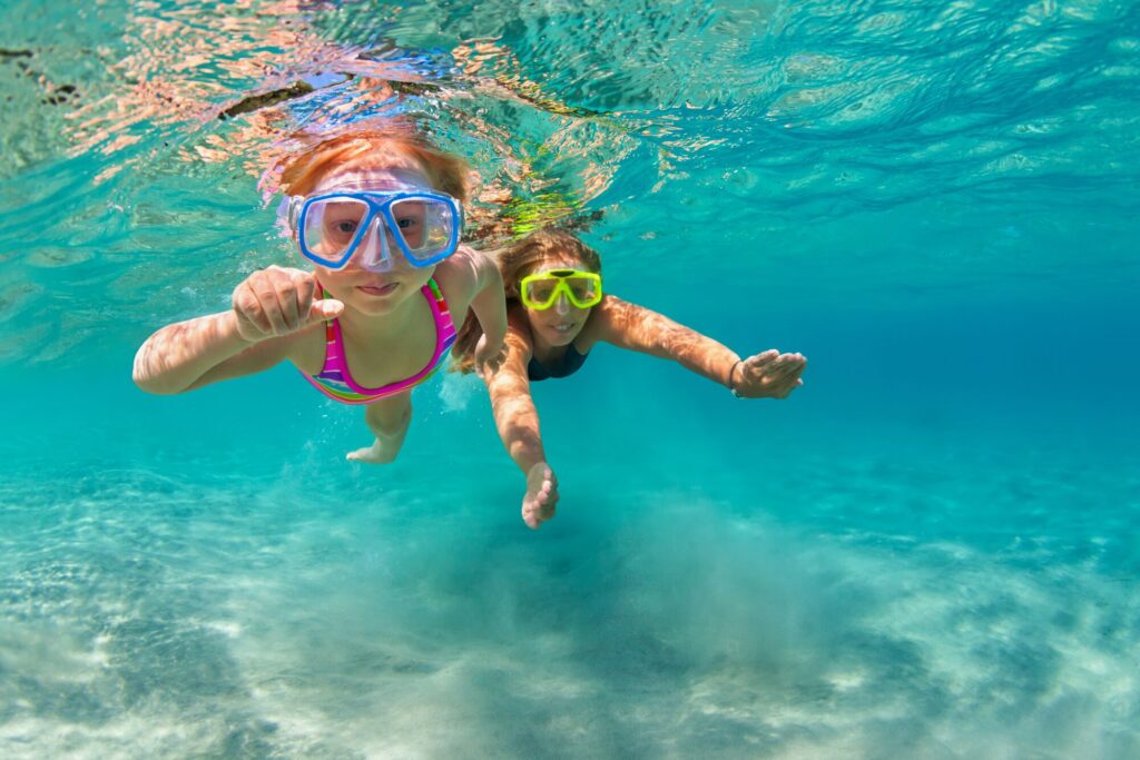 Two children swim underwater, wearing colorful snorkeling gear. Bright blue water and sandy seabed enhance the vibrant, playful atmosphere.